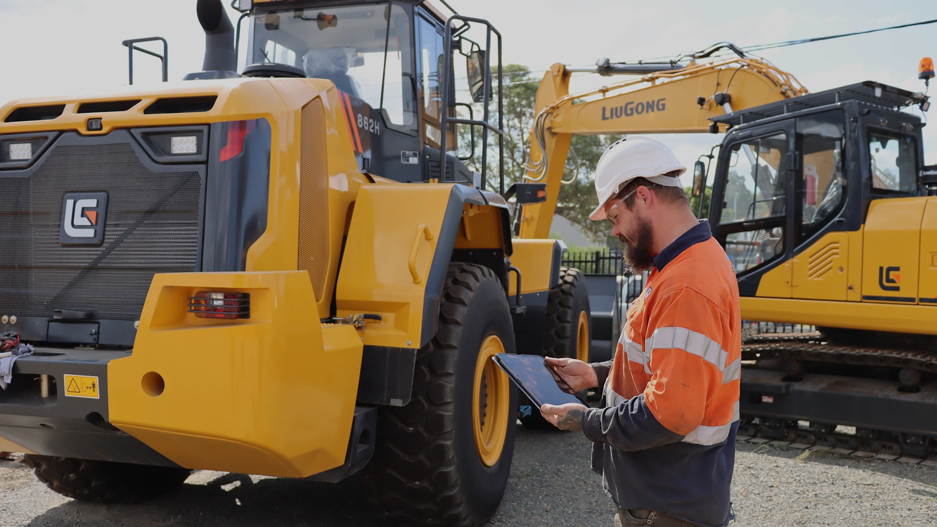 Man wearing orange hi vis looking at a device while standing in front of yellow heavy machinery