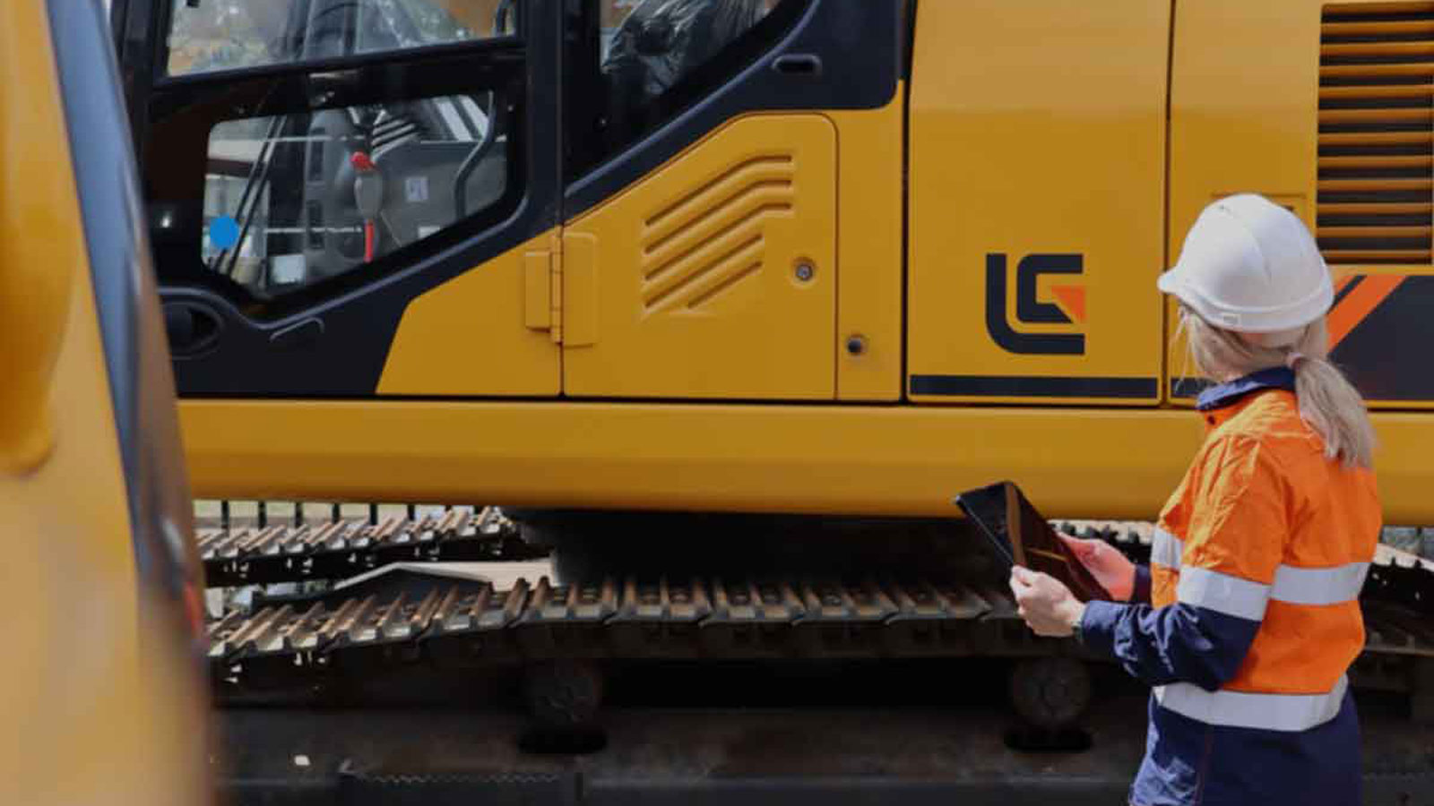 Girl in ppe with ipad doing risk assessment on a yellow excavator
