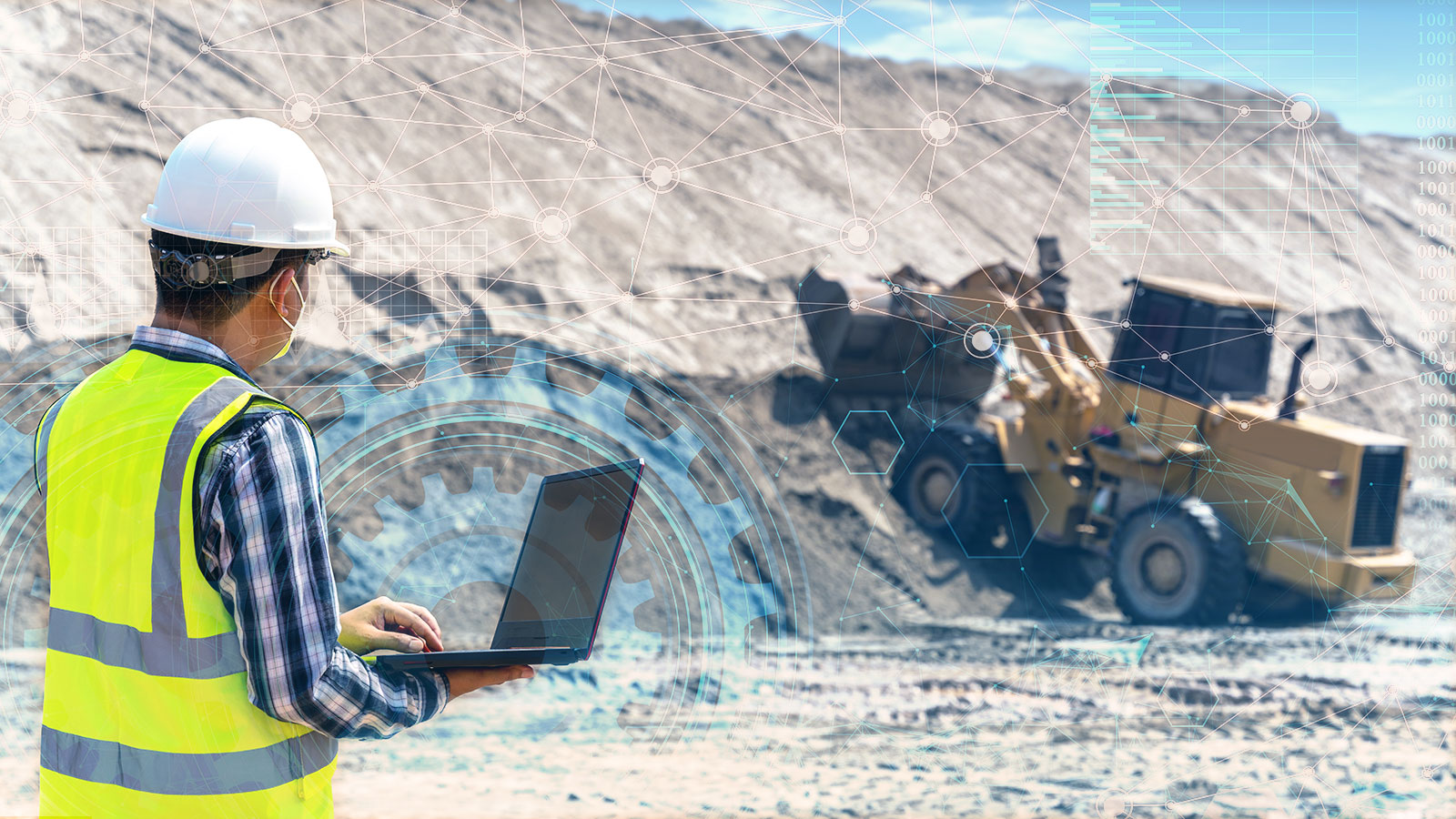 Person wearing PPE and holding a laptop inspecting yellow heavy machinery from afar