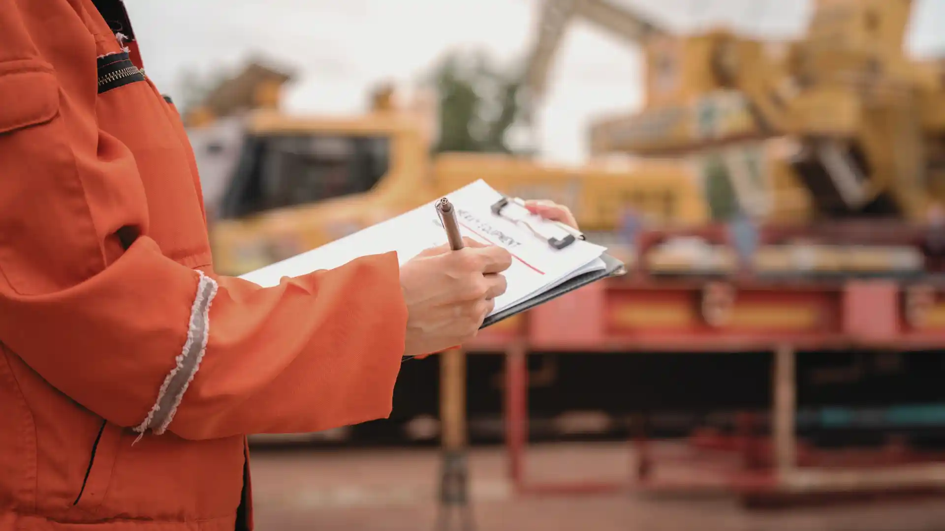 Person in hi-vis working on a clipboard while standing in front of yellow heavy machinery