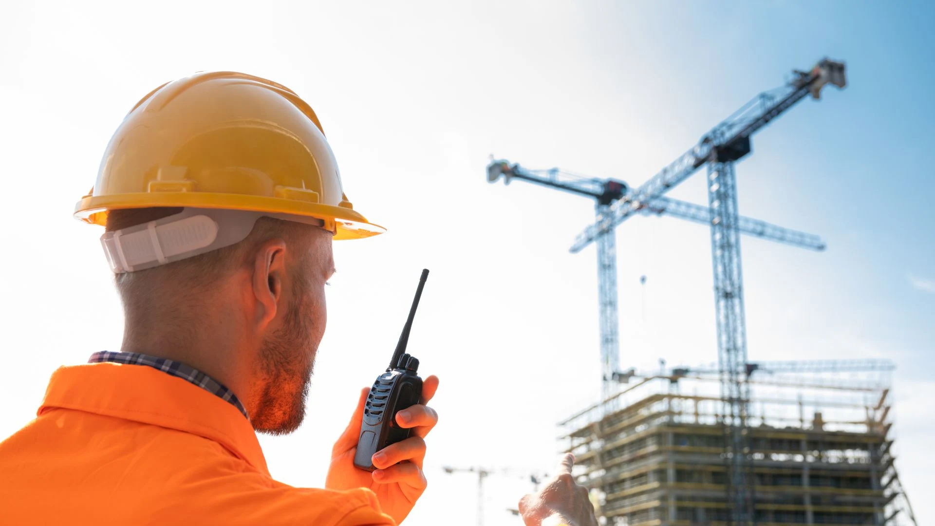 Man in PPE using a walkie talkie with a tall building being constructed by cranes in the background